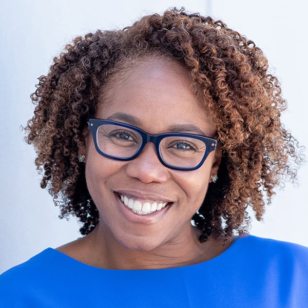 Headshot of Kahlilah Guyah, a black woman smiling with blue framed glasses, shoulder length curly brown hair, and a vibrant cobalt blue dress