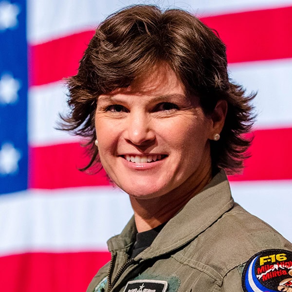 Headshot of Tracy LaTourrette, a white woman with layered brown shag haircut smiling in her fighter pilot uniform against an American flag